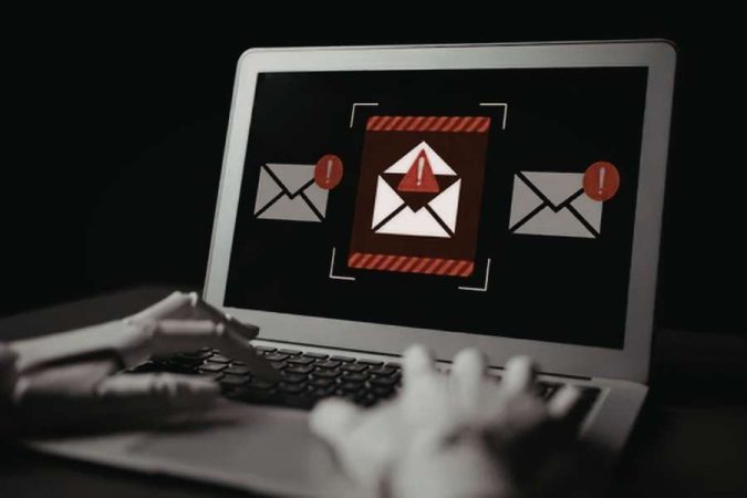 Des mains robotiques blanches tapent sur le clavier d'un ordinateur portable dont l'écran affiche une alerte de sécurité sur des e-mails suspects.