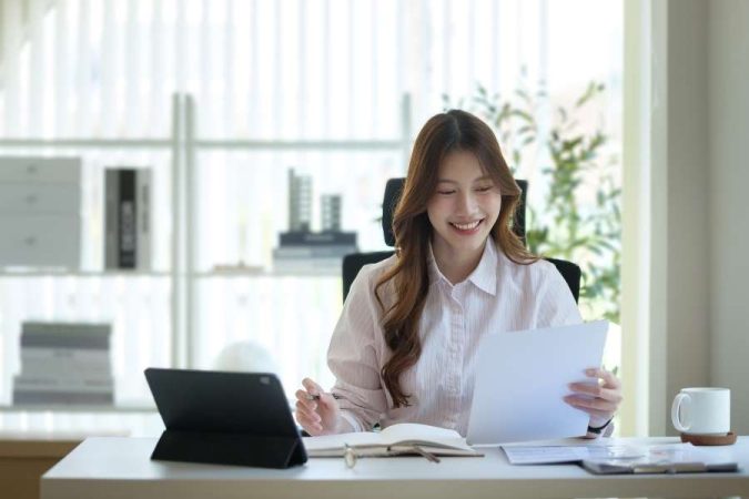Une jeune femme souriante examine des documents financiers assise à son bureau lumineux avec une tablette numérique.