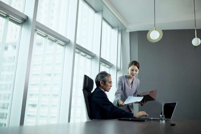 Une assistante professionnelle présente des documents imprimés à un cadre assis devant son ordinateur dans un bureau lumineux.