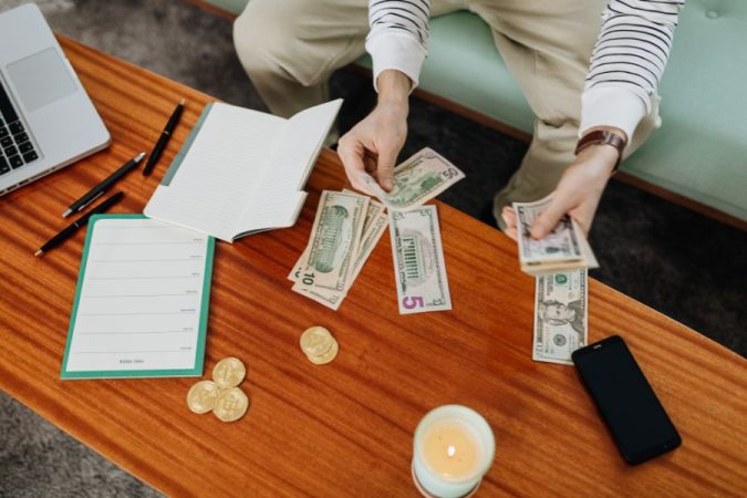 Une personne assise compte des billets en dollars sur une table en bois aux côtés d'un carnet de notes et d'un ordinateur, illustrant des astuces pour réduire ses dépenses.
